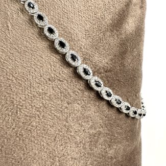 Colierul Colier Safir Black Diamonds, cu pietre prețioase întunecate în formă ovală, înconjurate de pietre limpezi strălucitoare, se întinde elegant pe un fundal moale de țesătură bej.